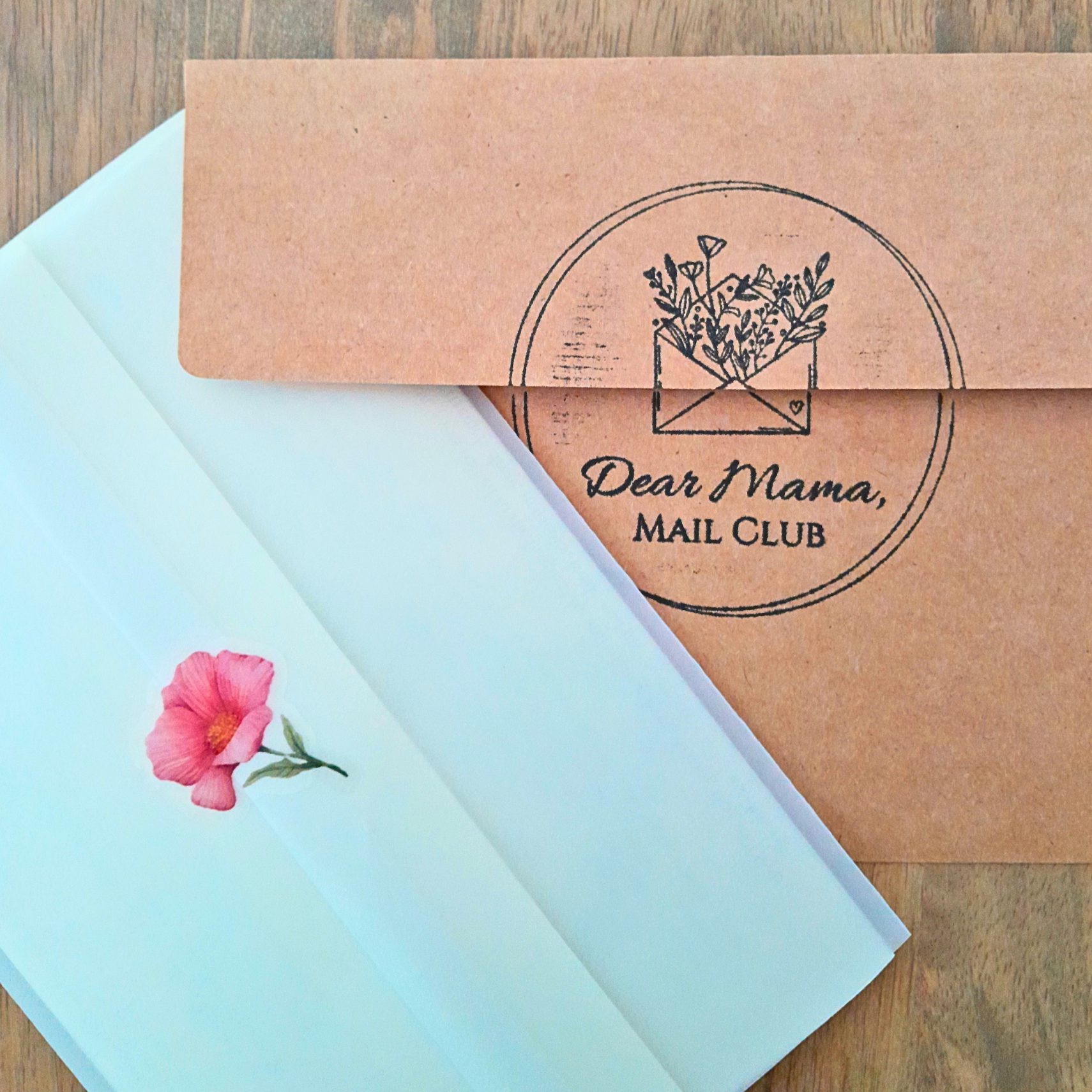 Kraft paper box with 'Dear Mama Mail Club' logo and a light blue card with a pink flower on a wooden surface.